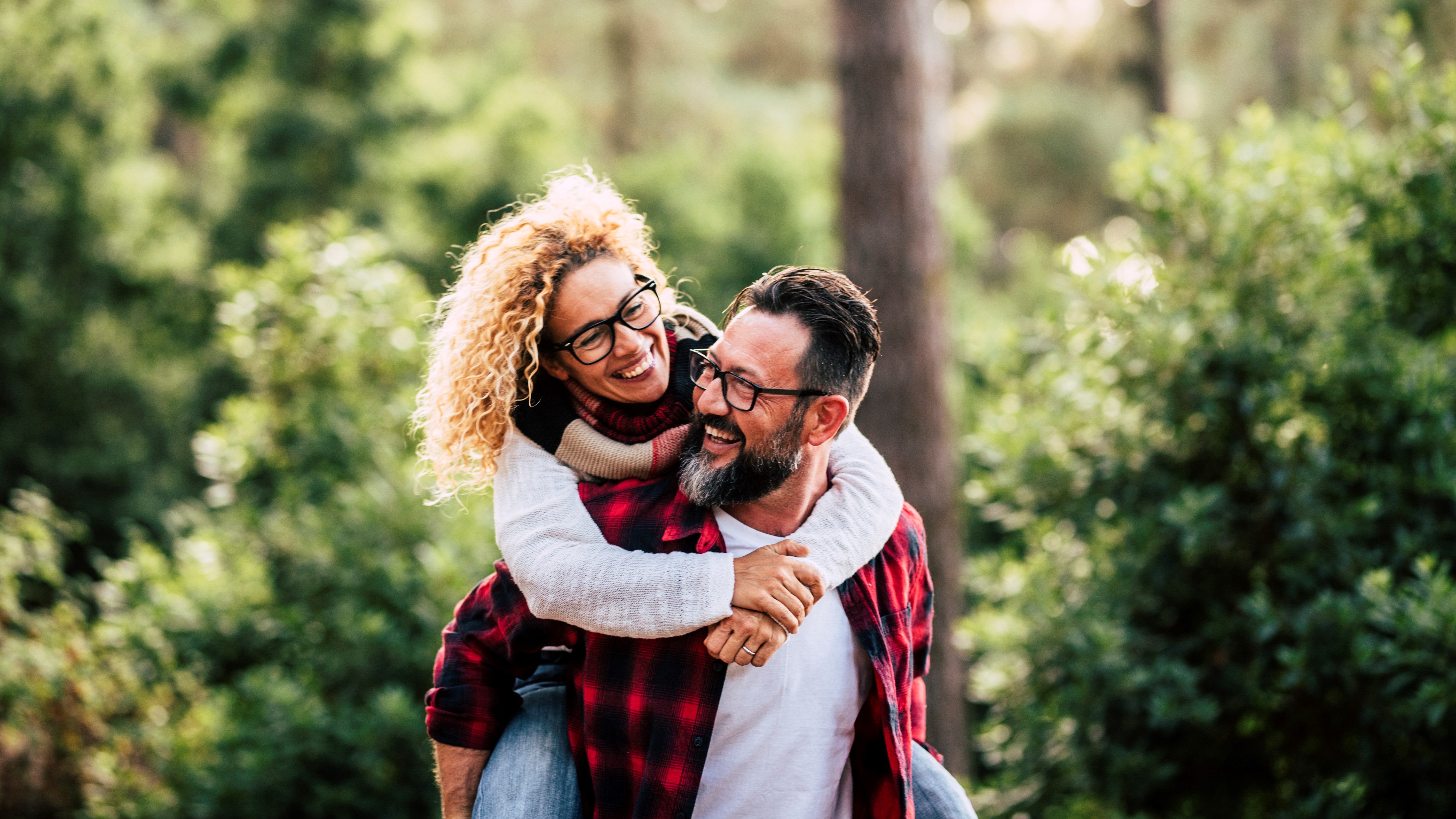 Older man with beard carrying a woman with curly hair, both smiling, on his back in the woods.