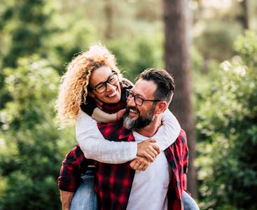Older man with beard carrying a woman with curly hair, both smiling, on his back in the woods.
