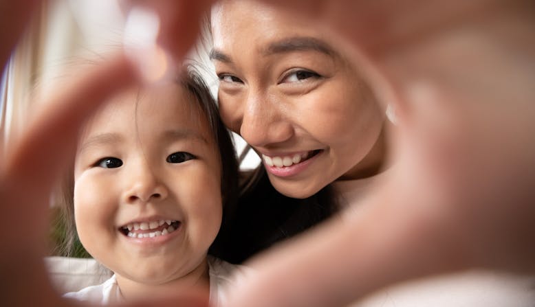 An adult and child smile while making the shape of a heart with their hands.
