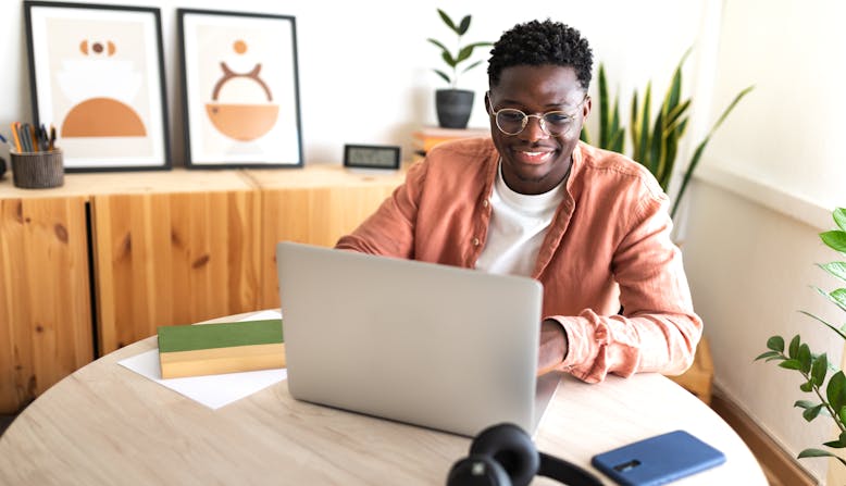 A Black man with glasses sits at a table at home working on his computer and smiling.