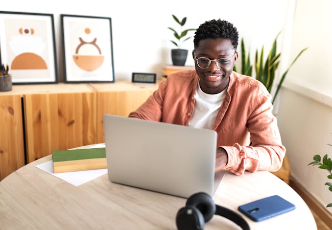 A Black man with glasses sits at a table at home working on his computer and smiling.