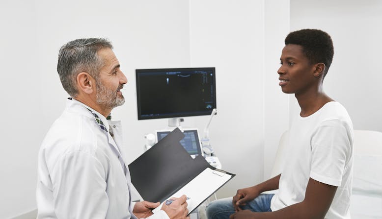 A doctor with light skin talking to a young male patient with skin of color.