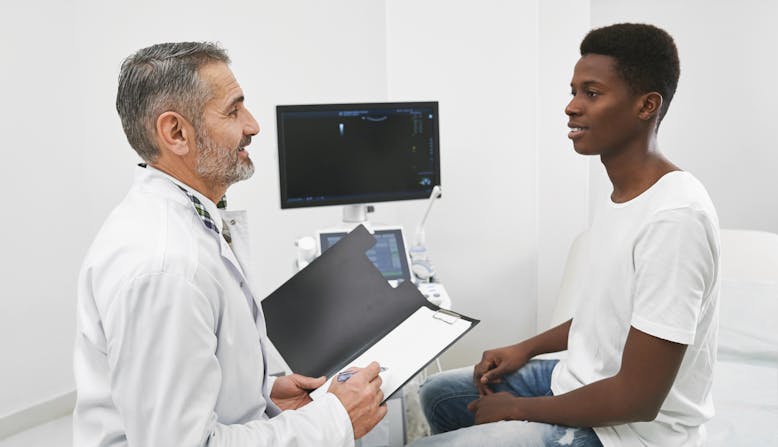 A doctor with light skin talking to a young male patient with skin of color.