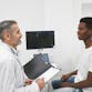 A doctor with light skin talking to a young male patient with skin of color.
