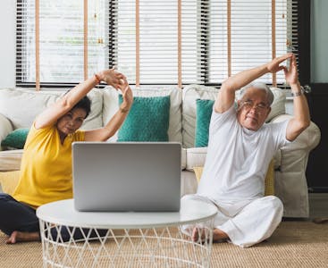 Two older couple stretching in front of a couch and laptop.