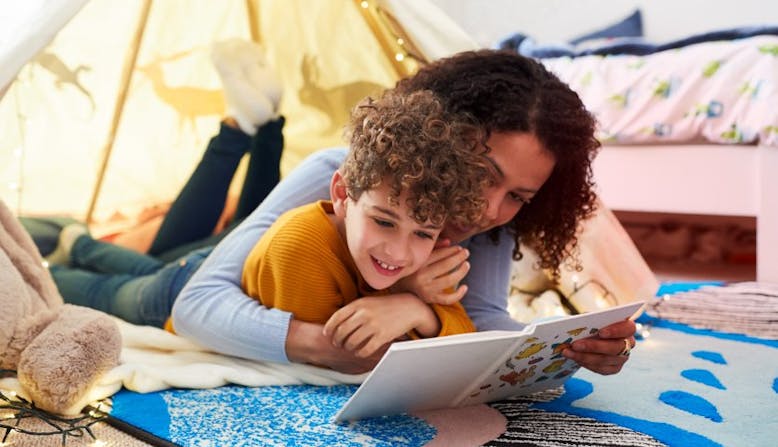 A mother and child lay in a tent and read a book together.