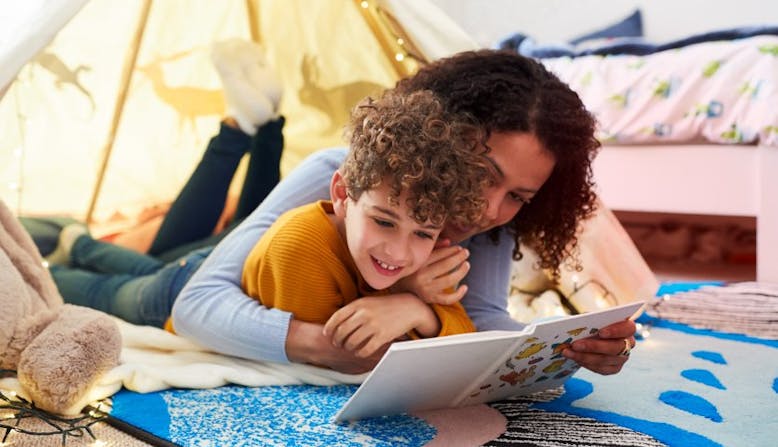 A mother and child lay in a tent and read a book together. 