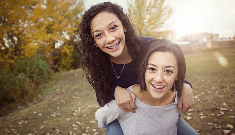 A girl with braces riding piggy back on her twin sister.