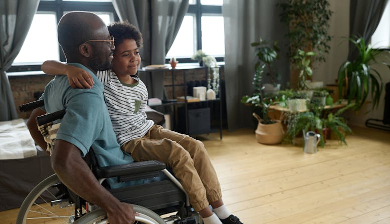 A middle-aged black man sits in a wheelchair with his son on his lap in their living room. 