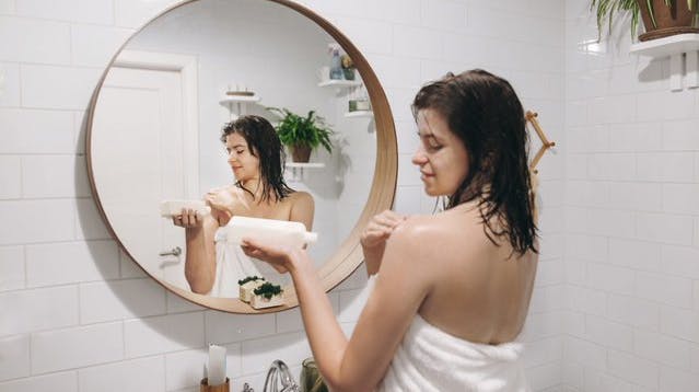 A woman in a towel holds a bottle and applies a cream to her shoulders in a bathroom. 