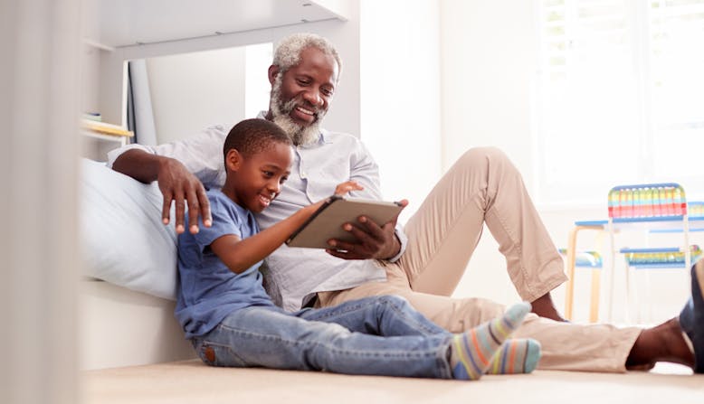 A grandfather sitting with his grandson in a bedroom using digital tablet together.