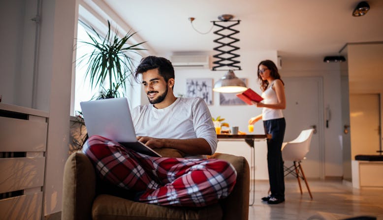 A man sits in his living room smiling and using his laptop with a roommate in the background.