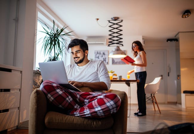 A man sits in his living room smiling and using his laptop with a roommate in the background.