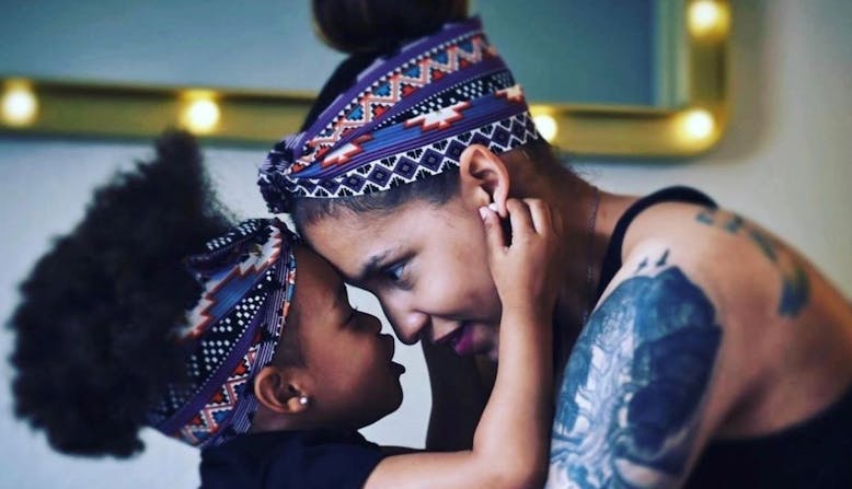 Mother and daughter touch foreheads, each wearing patterned headbands. The mother has tattoos on her arm and shoulder.