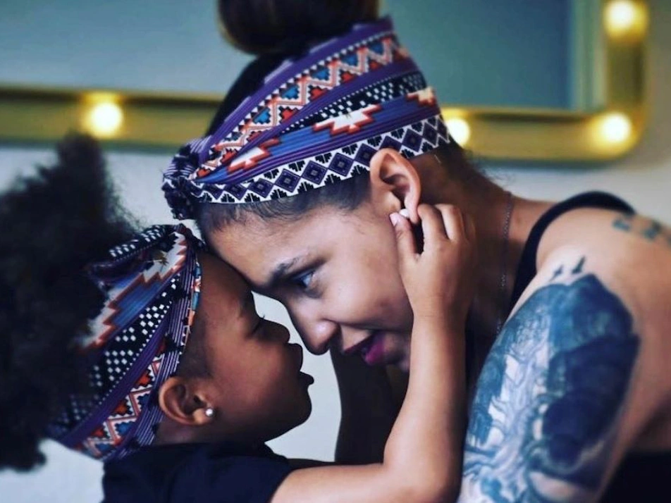 Mother and daughter touch foreheads, each wearing patterned headbands. The mother has tattoos on her arm and shoulder.