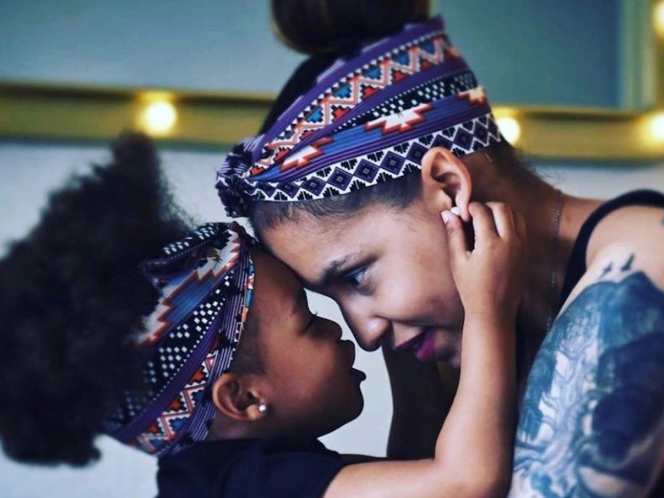 Mother and daughter touch foreheads, each wearing patterned headbands. The mother has tattoos on her arm and shoulder.