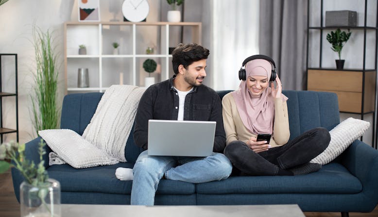 A couple sit on a couch in their living room, using a laptop and phone to watch and listen to media.