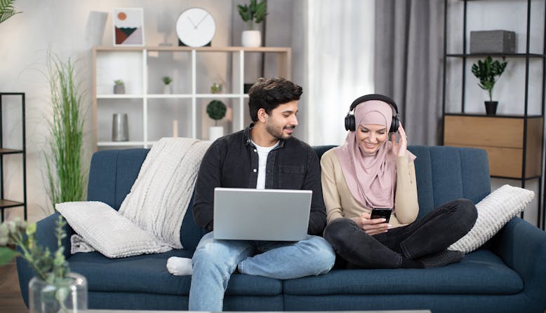 A couple sit on a couch in their living room, using a laptop and phone to watch and listen to media.