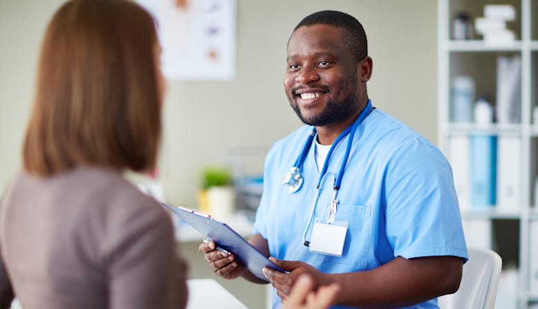 A black male nurse holds a clipboard and talks to a woman patient.