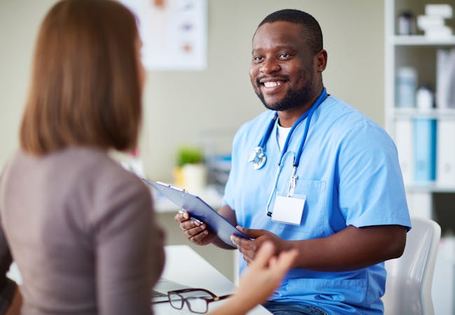 A black male nurse holds a clipboard and talks to a woman patient.