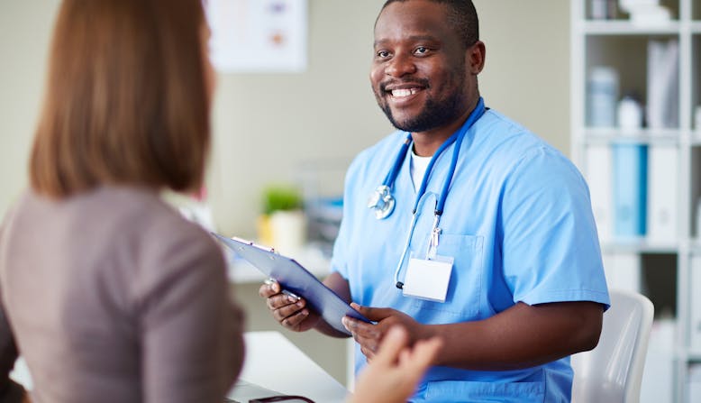 A black male nurse holds a clipboard and talks to a woman patient.