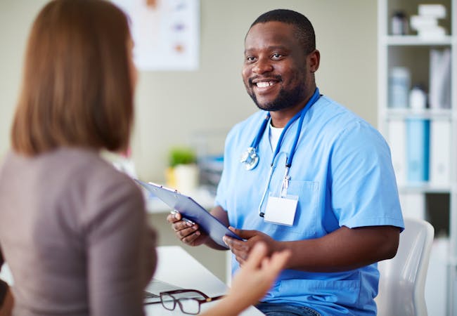 A black male nurse holds a clipboard and talks to a woman patient.