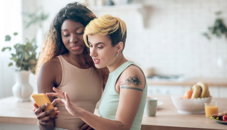 A woman with dark skin and a woman of lighter skin both look at a mobile phone.