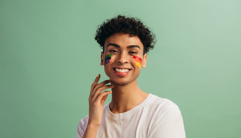 Young man with light brown skin and dark curly hair smiling with his hand near his face. His cheeks are painted in the pride LGBTQ flag.