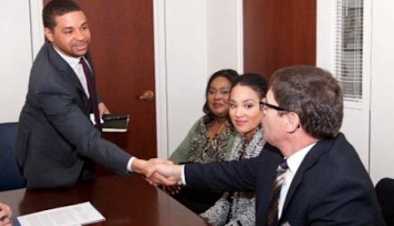 African American man standing and shaking hands with a white man who is sitting. Two African American women sit between and look on.