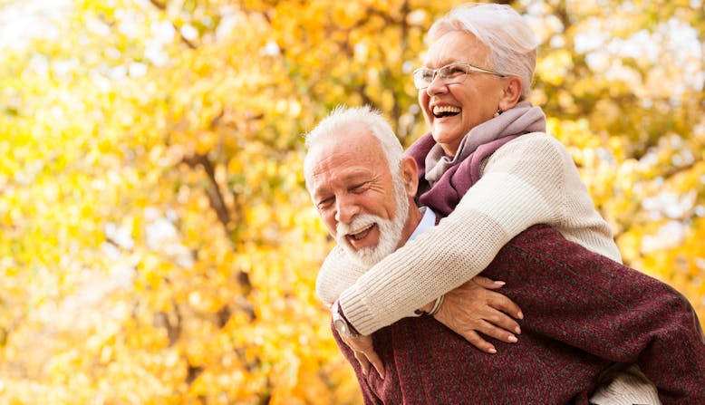 A couple walks in a park smiling with fall leaves in the background.