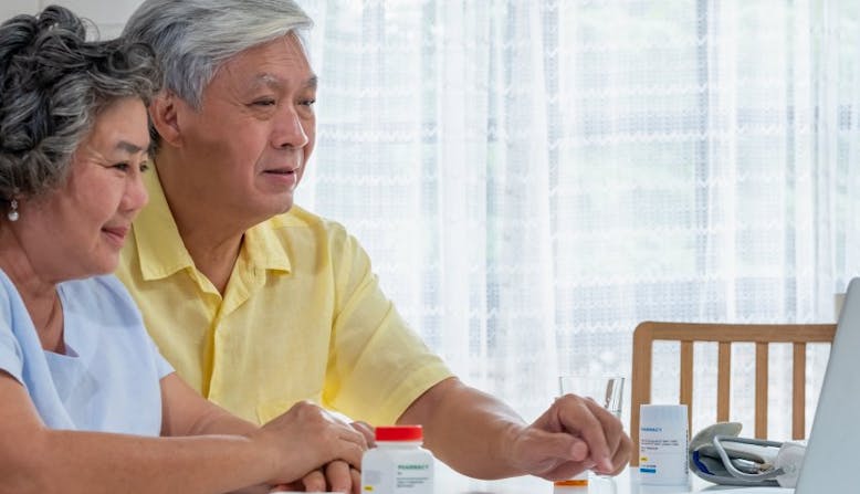 An older couple sits in front of a laptop and medication bottles. 
