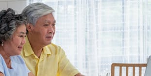 An older couple sits in front of a laptop and medication bottles.