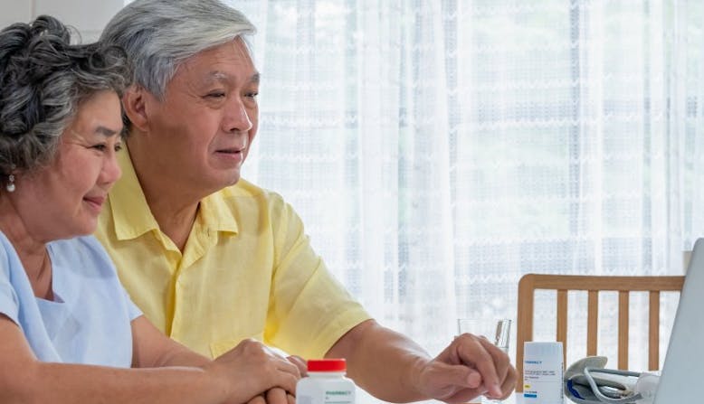An older couple sits in front of a laptop and medication bottles.