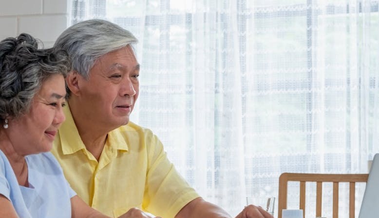 An older couple sits in front of a laptop and medication bottles.