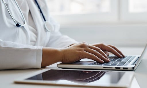 A doctor types on a laptop on a white desk. 