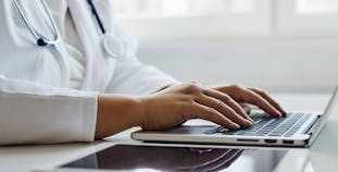 A doctor types on a laptop on a white desk. 