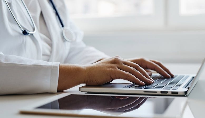 A doctor types on a laptop on a white desk. 