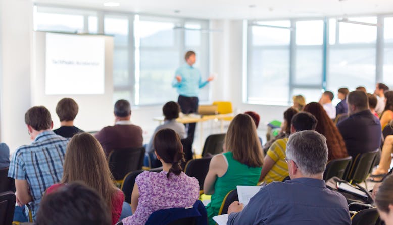 A group of people in chairs viewed from the back with a speaker presenting a slide show and the front of the room.