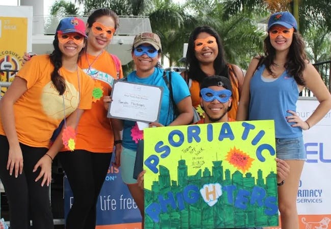 A group of people smiling and wearing superhero masks in NPF colors at an NPF event with a sign that says "Psoriatic Phighters".