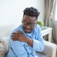 A black man sitting on a couch grips his shoulder in pain.