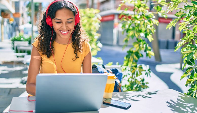 A young woman sits at a table outside with a laptop and headphones.