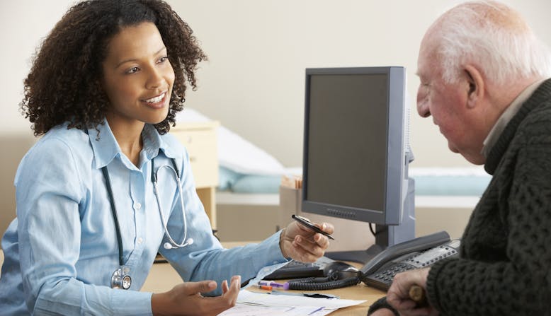 A man speaks to a doctor at her desk in front of a computer.
