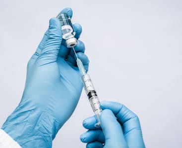Gloved hands holding a syringe inserted into a vial.