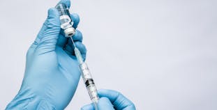 Gloved hands holding a syringe inserted into a vial.