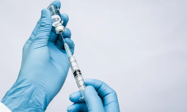 Gloved hands holding a syringe inserted into a vial.