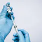 Gloved hands holding a syringe inserted into a vial.