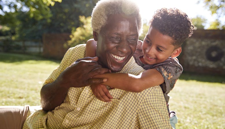 A smiling man sitting on grass, embraced by his grandson.