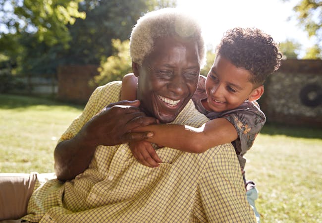 A smiling man sitting on grass, embraced by his grandson.
