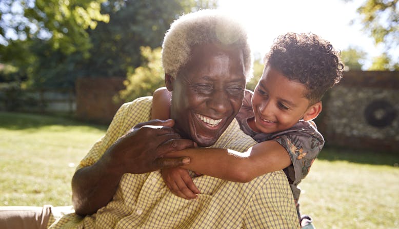 A smiling man sitting on grass, embraced by his grandson.