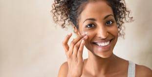 A woman smiles while putting lotion on her face.